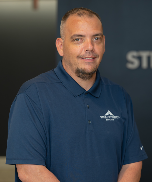 Man in blue shirt with Straightaway Tire & Auto logo, smiling indoors.