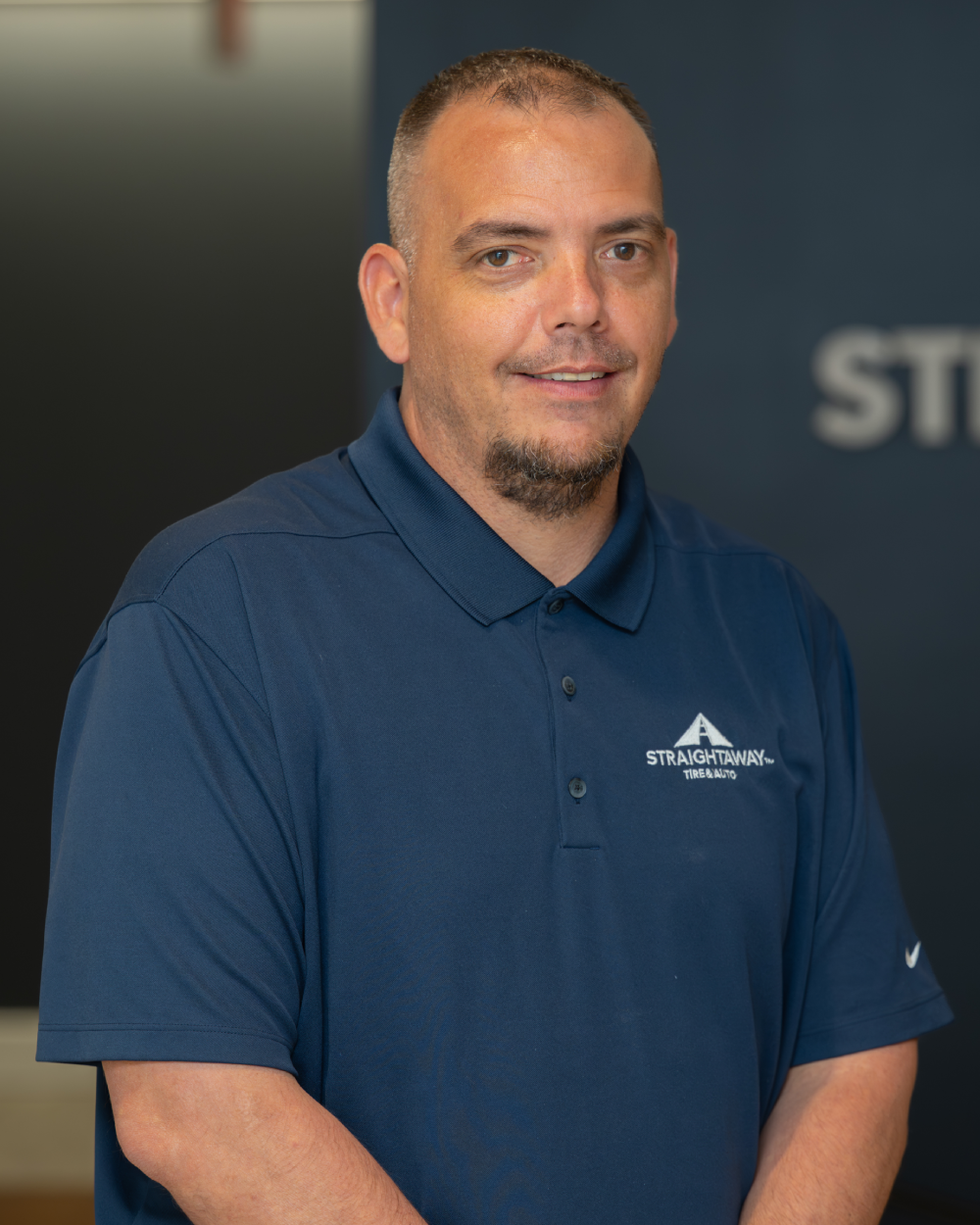 Man in blue shirt with Straightaway Tire & Auto logo, smiling indoors.