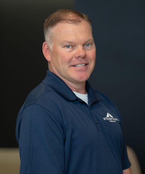 Man in blue shirt smiling, standing against a dark background.