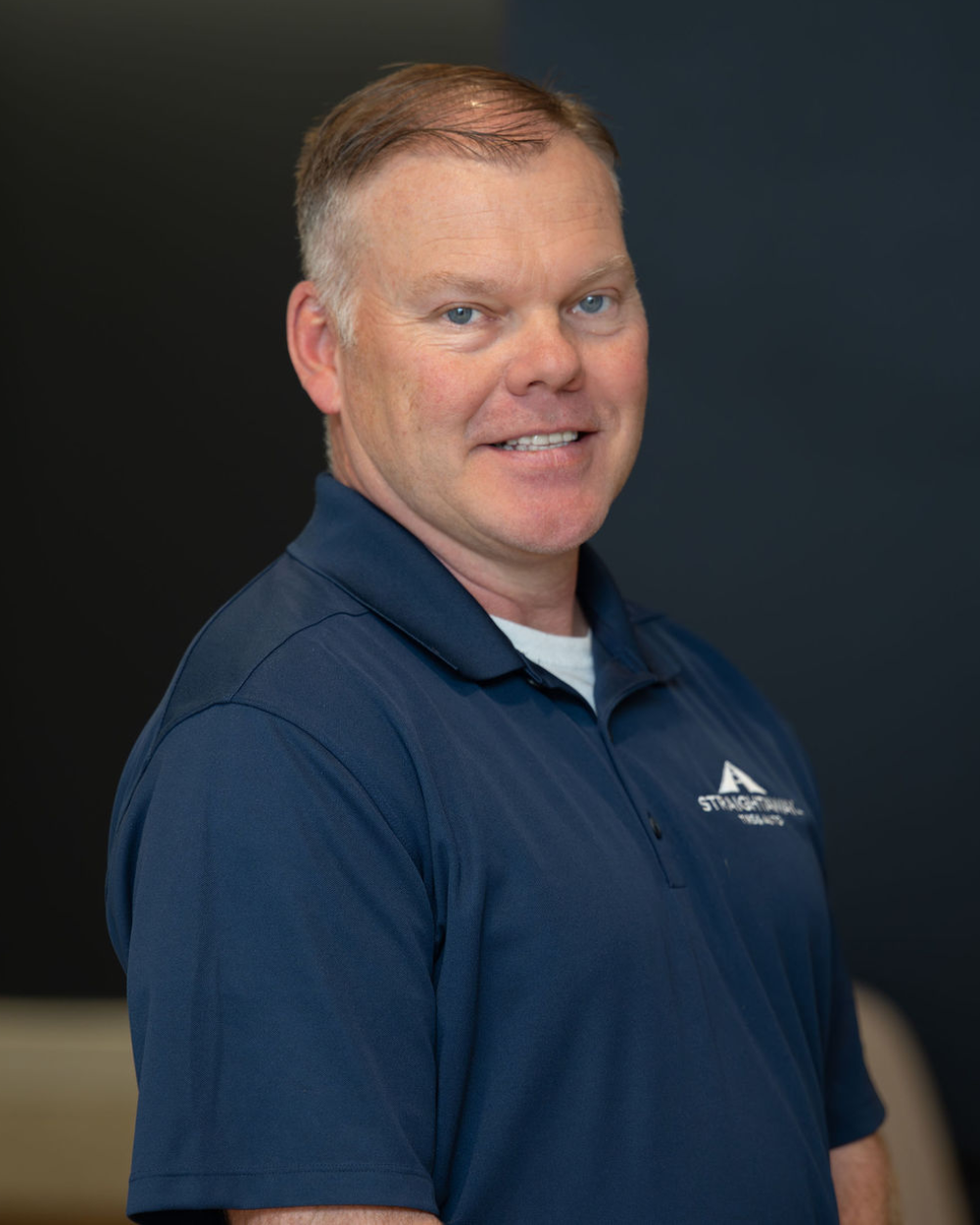 Man in blue shirt smiling, standing against a dark background.