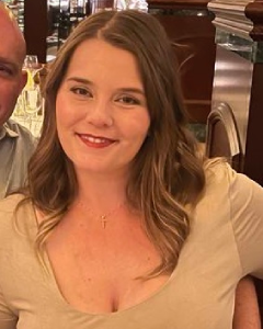 Smiling Woman In A Beige Top At A Restaurant, With A Blurred Male Figure Beside Her. Warm Lighting Sets A Cozy Atmosphere.