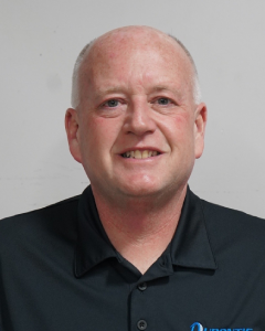 Smiling Man In A Black Collared Shirt Against A Neutral Background.