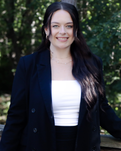 Smiling Woman With Long Dark Hair In A Black Blazer Stands Outdoors Against A Backdrop Of Green Trees.