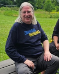 A Person With Long Gray Hair, Sitting Outdoors On A Bench, Wearing A Dark Blue Shirt With Yellow And Blue Text.