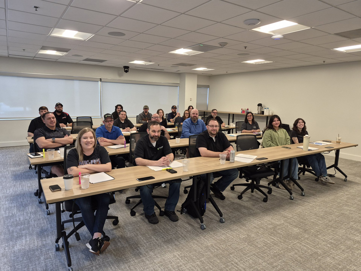 A group of people in a classroom setting, smiling and seated at desks, participating in a workshop or training session.
