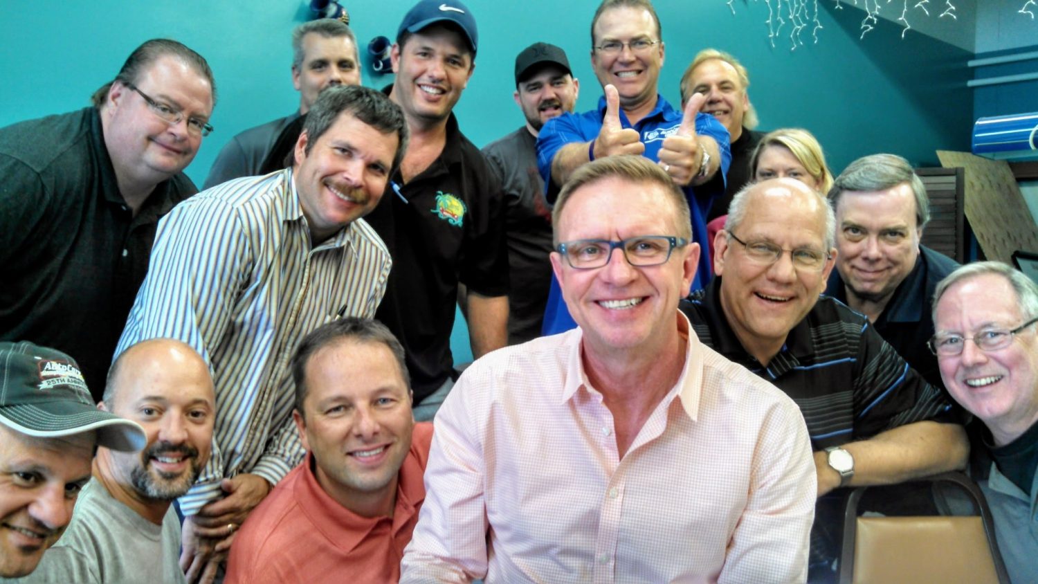 A group of smiling people at a casual office meeting with a teal background and festive string lights.