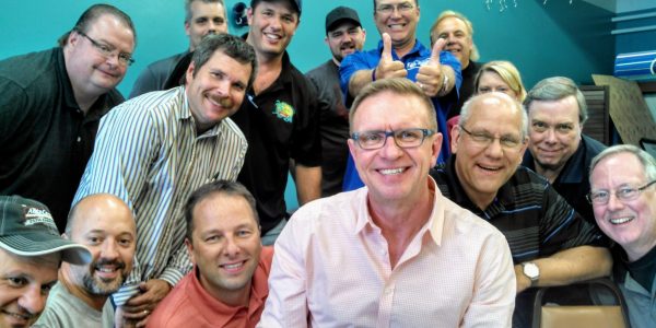 A group of smiling people at a casual office meeting with a teal background and festive string lights.